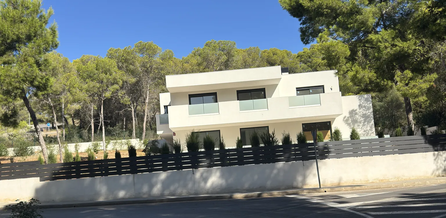 Modern white new build with glass balconies behind a fence, surrounded by pine trees in Calvià